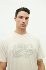 Remera de manga corta color beige con estampado gráfico frontal de la marca Brooksfield en tonos grisáceos, con diseño de estilo vintage y tipografía clásica.