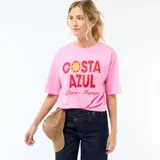 Camiseta de corte oversize color rosa, con cuello redondo y mangas cortas. Presenta un estampado frontal con la frase 'COSTA AZUL' en letras rojas y un diseño de sol, acompañado por la inscripción 'Palma de Majorque'.