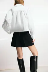 Camisa de cuello solapa y manga larga abullonada con puños abotonados. Presenta un diseño de corte recto con cierre frontal oculto por solapa.