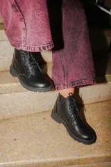 Bota tipo borcego de cuero negro, con cordones frontales, cierre lateral y suela track con costura blanca visible.