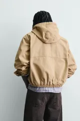 Cazadora relaxed fit de tejido de algodón con efecto lavado, color tostado. Cuenta con cuello con capucha, manga larga con puño abotonado, bolsillos de vivo en la cadera, bajo elástico y cierre frontal con alamares metálicos y cremallera oculta.