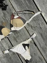 Conjunto de bikini con estampado abstracto en tonos tierra, blanco y negro. El top es de estilo triángulo fijo con breteles regulables y cierre de gancho en la espalda. La bombacha es de corte vedetina con laterales fruncidos elásticos.
