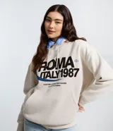 Buzo oversized color beige con capucha ajustable y bolsillo canguro. Presenta un estampado frontal estilo college con el texto "ROMA ITALY 1987" en negro y detalles en azul. Tiene puños y bajo ajustados.