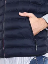 Chaleco acolchado de nailon en color azul marino, con cuello alto, cierre frontal de cremallera con detalles distintivos de la marca y bolsillos laterales con cierre. Presenta el logo de Tommy Hilfiger bordado en el pecho.