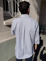 Camisa de corte clásico confeccionada en algodón, con diseño de rayas verticales finas en color rosa sobre fondo blanco. Presenta cuello abotonado, cierre frontal con botones, mangas largas con puños abotonados y un bolsillo en el pecho con el logo de la marca bordado.