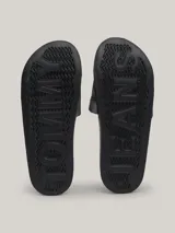 Ojotas tipo slide con base negra y pala ancha acolchada con acabado metalizado gris. Presentan un parche con el logo de Tommy Jeans en la parte superior de la pala.