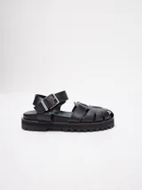 Sandalias romanas Mutma modelo Gena, color negro, confeccionadas en cuero vacuno wax. Presentan diseño de tiras cruzadas, tira ajustable al tobillo con hebilla metálica y suela de goma con plataforma track.