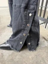 Pantalon de denim negro de corte recto y tiro alto, con aberturas laterales en los tobillos que incluyen cierre de botones a presión y ruedo deshilachado.