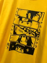 Remera amarilla con estampado de personajes de Naruto en recuadros negros con texto en japonés. El estampado incluye a Naruto, Sasuke, Sakura y Kakashi.
