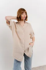 Camisa larga de lino color beige, con corte recto y calce suelto. Presenta cuello camisero, botones frontales y mangas que se pueden arremangar con presilla y botón.