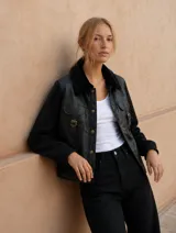 Campera de estilo utilitario con cuerpo de cuero sintético negro y mangas de tejido polar. Presenta cuello con solapas de piel sintética, bolsillos frontales con solapa y hebillas decorativas, y cierre frontal con botones a presión metálicos.