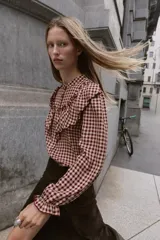 Blusa de manga larga con estampado de cuadros vichy en tonos burdeos y beige. Presenta cuello mao y grandes volantes fruncidos que cruzan el pecho y los hombros. Los puños son elásticos y fruncidos.