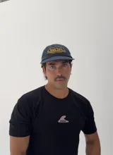 Gorro tipo gorra de béisbol color azul marino, con visera corta y diseño bordado en amarillo con la palabra "But'ton" y la frase "Cruise to Victory".