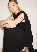 Vestido largo negro de corte entallado y diseño asimétrico, sin mangas, con detalle de capa en el cuello y fruncidos laterales.