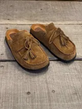 Zueco tipo mocasín destalonado confeccionado en gamuza color tabaco, con detalle de cordones y borlas en el empeine y costura reforzada en el contorno de la puntera.