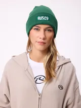 Gorro tipo beanie de tejido de punto acanalado en color marrón, con parche rectangular frontal con el logo de la marca Rusty.