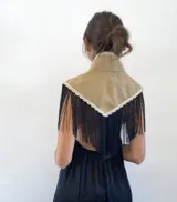 Hombrera de cuello alto con diseño triangular en la espalda, confeccionada en tejido de textura rústica color tostado, con borde de encaje blanco y flecos largos negros.