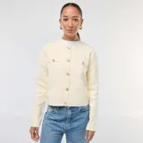 Cárdigan de punto texturizado en color beige, con cuello redondo, manga larga y cierre frontal mediante botones dorados. Presenta dos bolsillos decorativos en el pecho con botones a juego.