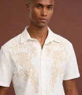 Camisa de corte comfort confeccionada en algodón, color blanco, con cuello solapa, manga corta y cierre frontal de botones. Presenta un delicado bordado floral en tono beige en la parte superior delantera.