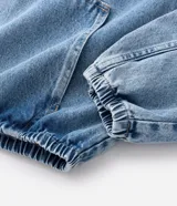 Campera tipo bomber confeccionada en tejido de jean, con cierre frontal de cremallera, capucha ajustable con cordones y bolsillos frontales. Presenta puños y cintura elásticos, ofreciendo un ajuste cómodo y un estilo urbano.