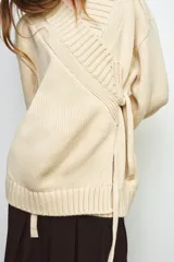 Chaqueta cruzada de punto color beige con manga larga, cuello smoking y cierre frontal con lazada. Presenta detalles de calados en los puños.
