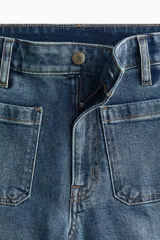 Jean de tiro alto con corte wide leg, confeccionado en denim de algodón con leve elasticidad. Presenta bolsillos de parche frontales, cierre con botón y costuras marcadas a lo largo de las piernas.