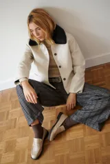 Chaqueta corta color beige con cuello de piel sintética negra, cierre frontal de botones y bolsillos laterales.