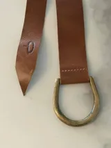 Cinto de cuero flexible diseñado para anudar, con terminación en punta y detalle de marca grabada en el extremo. Incluye una hebilla metálica en forma de D en tono bronce.