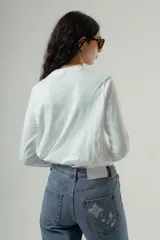 Camiseta de manga larga, cuello redondo, con rayas horizontales marrones sobre fondo blanco. Presenta un pequeño logo bordado en rojo en el pecho.