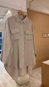 Camisa color beige de lino con botones de nácar, manga larga, dos bolsillos delanteros con solapa, más larga en la parte trasera que en la delantera y con cortes en los costados.