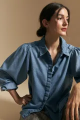 Camisa de denim azul con cuello camisero, cierre frontal de botones y mangas largas abullonadas con hombros fruncidos.
