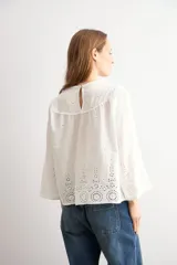 Blusa blanca de algodón con diseño de encaje bordado tipo broderie en el cuello, mangas y ruedo. Presenta mangas tres cuartos acampanadas y un corte holgado.