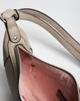 Bandolera de diseño semicircular en color taupe, confeccionada en símil cuero. Presenta detalles de tachas metálicas plateadas en los bordes, placa con logo de la marca en el frente y correa ajustable para hombro o cruzada.