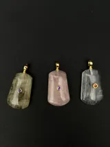Dije de piedra natural con terminación pulida y detalle de piedra pequeña engarzada en metal dorado. Presenta un diseño rectangular con bordes redondeados y argolla superior para cadena.