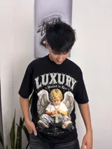 Remera negra de corte oversize con estampado frontal de estilo barroco/urbano que incluye un ángel con alas, cadenas, una cruz y la inscripción "Luxury".