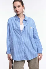 Camisa oversize de popelina a rayas celestes y blancas, con cuello camisero, manga larga con puños abotonados, cierre frontal con botones y un bolsillo en el pecho con etiqueta bordada.