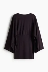 Vestido corto de punto fino en color negro, con escote bote, espalda abierta y mangas capa largas con diseño abullonado y costura fruncida en la cintura.