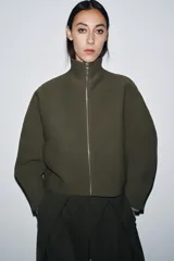 Chaqueta corta de tejido grueso color verde oliva, con cuello alto y cierre frontal completo de cremallera plateada. Presenta un corte amplio y mangas largas caídas.