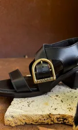 Sandalias de cuero color marrón con tiras anchas cruzadas y hebilla metálica decorativa en forma de estribo. Presentan taco cuadrado de altura media y suela de goma.