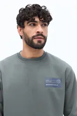 Buzo de algodón y poliéster color verde musgo, con cuello redondo y puños acanalados. Presenta un parche estampado rectangular con texto y un logo circular en el pecho.