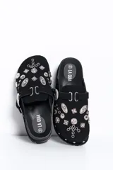 Zueco negro de estilo slip-on, confeccionado con acabado similar a gamuza, suela gruesa dentada de goma, hebilla lateral plateada de diseño occidental y apliques metálicos decorativos con tachas y ojetes en la capellada.