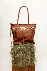 Cartera de cuero color camel, de silueta amplia y blanda, con manijas largas y terminación artesanal en los bordes. Cuenta con apliques decorativos frontales de alpaca y nácar, tientos largos colgantes y bolsillo interno.