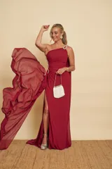 Vestido largo de fiesta color bordó, de un solo hombro, con detalles de pedrería en el tirante y fruncidos laterales. Cuenta con tajo pronunciado en la pierna y una capa larga vaporosa que cae desde el hombro.