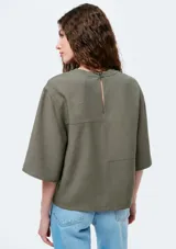 Blusa de manga corta color verde oliva, con cuello redondo y diseño de paneles con costuras visibles. Presenta un corte holgado y mangas con caída amplia.