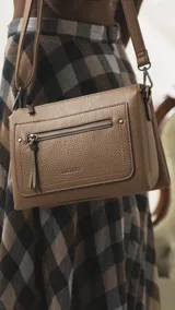 Bandolera de tamaño mediano en color gris, confeccionada en símil cuero con textura granulada. Presenta un bolsillo frontal con cierre, herrajes metálicos oscuros y correa ajustable y desmontable.