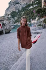 Camisa bordó de cuello alto ajustable con doble hebilla y cierre frontal con botones ocultos por solapa. Tiene mangas cortas con trabillas y diseño holgado.