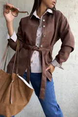 Sobrecamisa de gamuza color chocolate, con cierre frontal de botones metálicos y lazo ajustable en la cintura. Presenta cuello clásico y mangas largas con puños rectos.