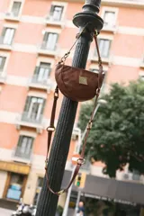 Cartera pequeña de cuero marrón con diseño semicircular, detalles metálicos de bronce y asa ajustable con ganchos.
