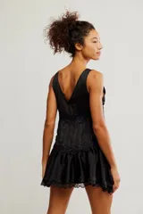 Mini vestido tipo slip de tela satinada negra, con escote en V profundo, diseño en capas con volados y detalles de encaje en los bordes. Corte estilo babydoll, sin mangas y silueta amplia.