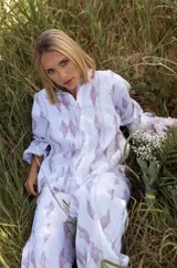 Camisa de lino de corte holgado, con cuello clásico, cierre frontal de botones y mangas largas. Presenta un estampado lineal en tonos violetas sobre fondo blanco.